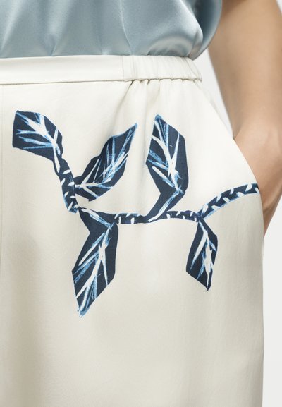Gros plan d'une jupe blanche avec un imprimé abstrait de feuilles bleu et noir, portée avec un haut bleu clair, montrant une taille élastique et une main dans la poche.