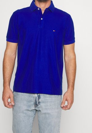 Niebieska koszulka polo wykonana z bawełny, z kołnierzykiem na guziki, krótkimi rękawami i małym czerwonym logo na lewej piersi. Noszona z jasnymi jeansami.