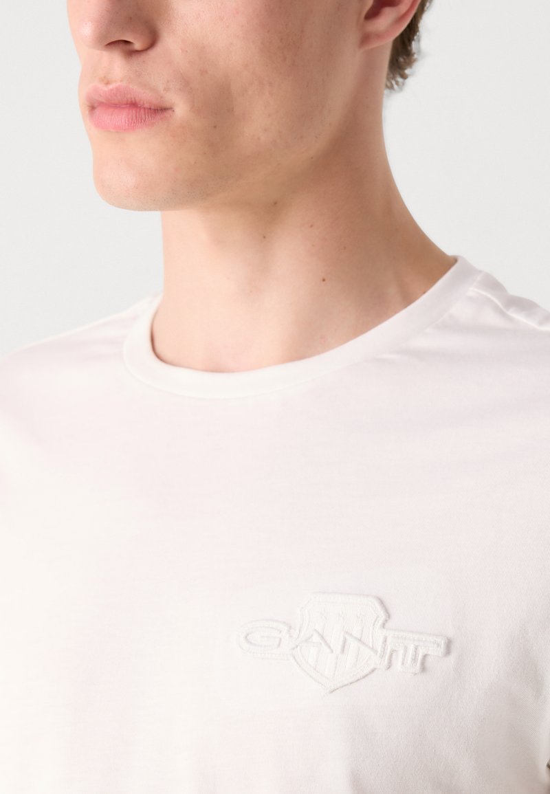 Weißes T-Shirt mit kurzen Ärmeln, das ein strukturiertes, gesticktes Logo auf der linken Brust zeigt. Das Material wirkt glatt und hat ein Rundhalsausschnitt-Design.