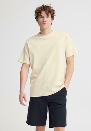 Junger Mann mit lockigem Haar, der ein beigefarbenes kurzärmeliges T-Shirt und schwarze Shorts trägt und mit einer Hand in der Tasche vor einem weißen Hintergrund steht.