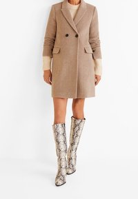 Femme portant un manteau beige croisé sur un pull crème avec des bottes hautes à motif peau de serpent, debout devant un fond blanc.