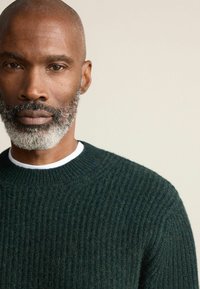 Dunkelgrüner, gerippter Strickpullover mit rundem Halsausschnitt, über einem weißen Hemd getragen. Die Textur wirkt weich und leicht flauschig.