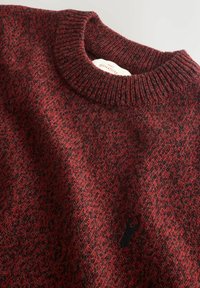 Close-up of a red and black knit sweater with a ribbed crew neck and small black deer logo on the chest.
