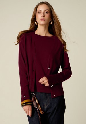 Femme aux longs cheveux ondulés porte un cardigan bordeaux boutonné sur un haut assorti et un pantalon foncé, tenant des porte-clés et portant des bracelets épais.