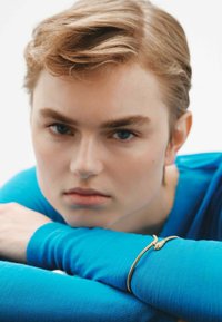 Top azul de mangas largas con tejido texturizado, que presenta una pulsera en forma de serpiente dorada en una muñeca. Primer plano de un rostro con expresión neutral.