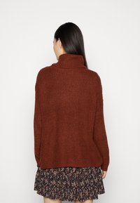 Rust-colored chunky knit sweater with a high collar, paired with a black floral tiered skirt, displayed from the back.