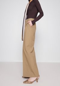 Kvinde i højtaljede beige wide-leg bukser, mørk tætsiddende langærmet bluse, m�ønstret tørklæde og slingback højhælede sko i slangeskind, stående sidelæns.