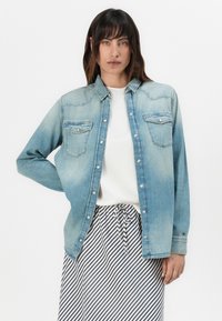 Camisa de ganga em azul claro, com colarinho clássico, dois bolsos no peito e fecho com botões. Usada sobre uma blusa branca, combinada com uma saia riscada.