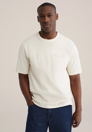 Roomkleurig katoenen T-shirt met korte mouwen, ronde hals en linkerborstzak, gecombineerd met donkerblauwe denim jeans.