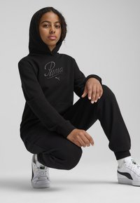 Haut à capuche noir avec un discret brodé "Puma", pantalon de survêtement noir assorti et baskets blanches avec des accents verts et argentés, présentant une silhouette classique.