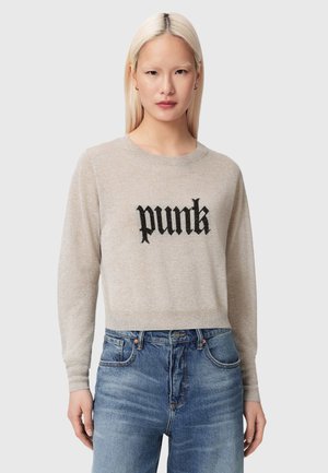 Γυναίκα με μακριά ξανθά μαλλιά που φοράει μπεζ πουλόβερ με την επιγραφή «punk» και μπλε τζιν, στέκεται μπροστά σε απλό γκρι φόντο.