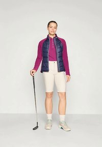 Jūras spēka pūka veste virs bordo krāsas garām piedurknēm polo krekla, kopā ar gaiši beidžiem šortiem. Uz kājām golfa apavi, rokā turot golfa nūju.