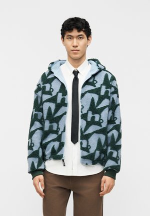 Noor mees, kes kannab sinise ja rohelise mustriga fliisjopet valge särgi, musta lipsu ja pruunide pükstega, seistes valge tausta ees.