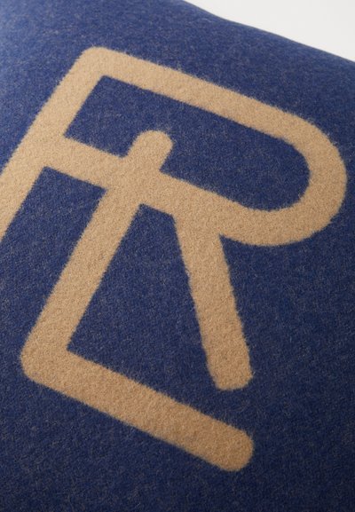 Blauwe vilten kussen met een groot, lichtbruin geometrisch letterontwerp. Zachte textuur met een gladde afwerking en zichtbare vezels.