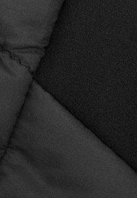 Schwarzer Stoff mit einer Mischung aus glatten und strukturierten Materialien, versehen mit einem gesteppten Muster, das die markanten Nähte und ein mattes Finish hervorhebt.