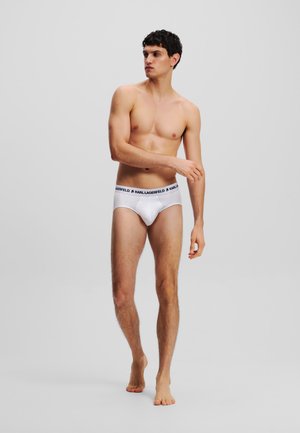 White brief undergarment with a black logo waistband, smooth material, fitted design, and mid-rise cut; model poses barefoot.