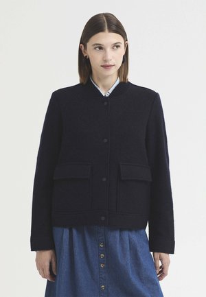 Jeune femme aux cheveux bruns et lisses porte une veste bleu marine boutonnée sur une jupe bleue boutonnée, debout devant un fond clair uni.