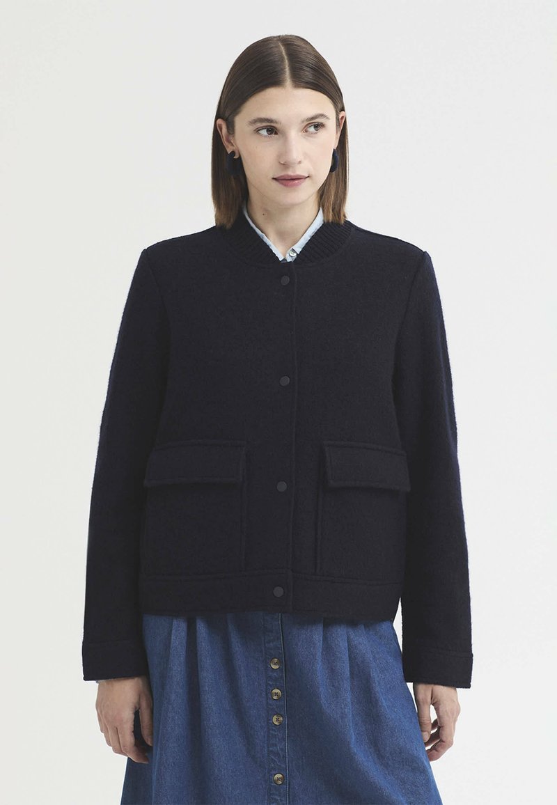 Jeune femme aux cheveux bruns et lisses porte une veste bleu marine boutonnée sur une jupe bleue boutonnée, debout devant un fond clair uni.