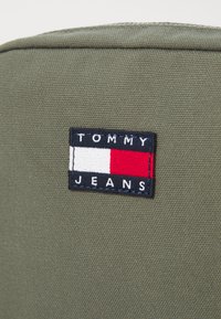 Il tessuto in tela verde presenta una toppa rettangolare con la scritta "TOMMY JEANS" e un motivo a strisce orizzontali rossa, bianca e blu.