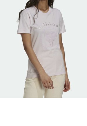 Kobieta w jasnoróżowej koszulce Adidas z haftowanym logo i dwoma rakietami tenisowymi, w połączeniu z jasnymi spodniami.