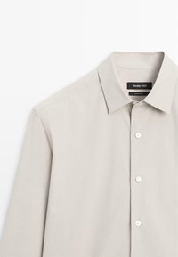 Chemise longue beige à manches longues avec col pointu et boutons blancs, étiquetée Massimo Dutti, fabriquée en Turquie.