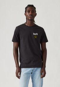 Mand med fletninger iført sort Levi's t-shirt med grøn grafik og lyseblå jeans, stående mod en ensfarvet lys baggrund.