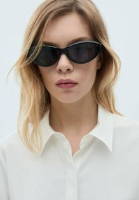 Schwarze, eckige Sonnenbrille mit dunklen Gläsern. Das Gestell hat eine glänzende Oberfläche und wird mit einem weißen Hemd getragen. Helles, welliges Haar ist sichtbar.