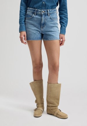 Γυναίκα που φοράει μπλε τζιν σορτς με ψηλή μέση, μακρυμάνικο τζιν πουκάμισο και μπεζ μπότες μέχρι τη μέση της γάμπας με αγκράφες, στέκεται σε λευκό φόντο.