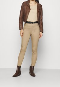 Brun läderjacka i kort modell över en beige topp, kombinerad med beiga åtsittande byxor och bruna ankelskortstövlar, med ett svart bälte med en metallspänne.