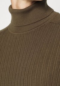 Maglione a collo alto marrone a coste con colletto alto e motivo a maglia testurizzata. Il tessuto appare morbido e flessibile. I dettagli si concentrano sulla scollatura.