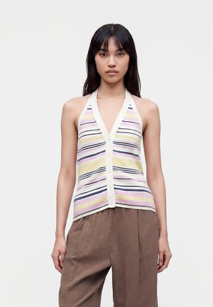 Femme portant un haut sans manches rayé à boutons dans des couleurs pastel et un pantalon marron ample, debout devant un fond uni.