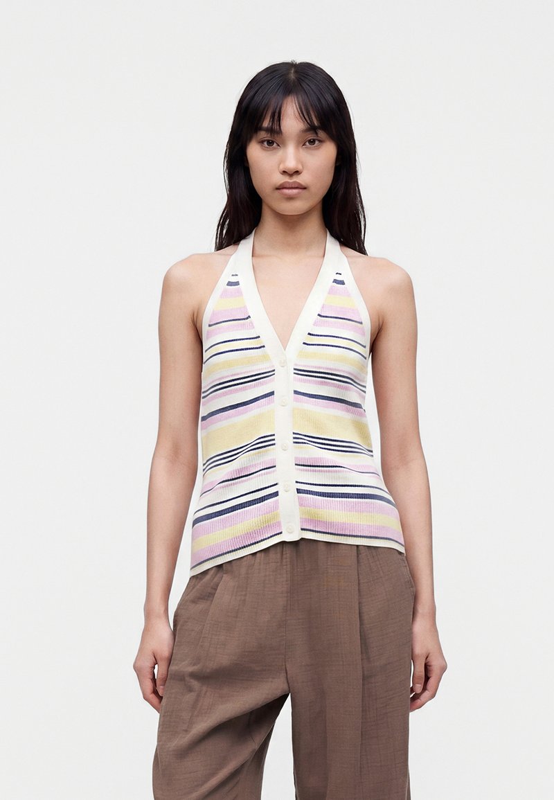 Femme portant un haut sans manches rayé à boutons dans des couleurs pastel et un pantalon marron ample, debout devant un fond uni.