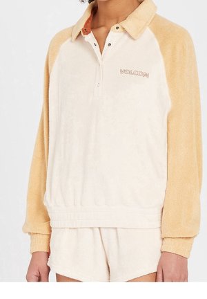 Pull-over en polaire à manches longues avec manches couleur beige et corps crème, boutons-pression au col, logo "VOLCOM" sur la poitrine, porté avec un short crème assorti.
