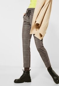 Pantalon à carreaux marron avec taille à cordon et poches latérales, associé à des bottes noires à lacets et un cardigan tricoté beige.