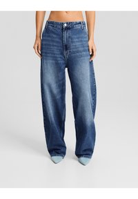 Jean en denim bleu à taille haute et jambes larges, avec une fermeture à boutons et une texture légèrement délavée ; stylisé avec des chaussures pointues bleu clair.