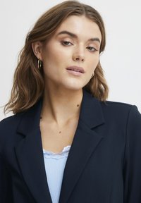 Jūras mužiks ar uzvalka atloka virs viegls zila ribotā topa ar mežģīņu apdari. Modelis ir ar viļņainiem matiem un maziem zelta auskariem gredzena formā.