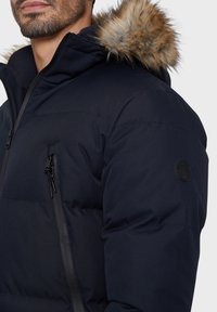 Marineblauwe pufferjas met een ritssluiting aan de voorkant, een borstzak en een afneembare rand van nepbont aan de capuchon. Structuurstof met een gladde afwerking.