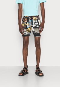 Shorts de bain noirs ornés d'un imprimé collage d'images variées, avec une taille élastique et un cordon de serrage. Portés avec des sandales noires et une chemise bleu clair.