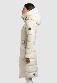 Beiger Steppmantel mit flauschigem weißem Kragen, tailliertem Bund und seitlichen Taschen, versehen mit einem Logo-Patch am Ärmel und gesteppter Oberfläche.