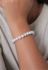 Poignet portant un bracelet de perles avec une seule perle ornée de strass, reposant sur un fond en tissu beige.
