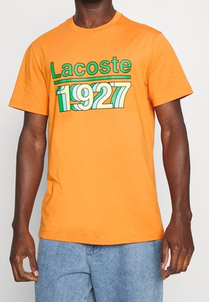 Homme portant un t-shirt orange Lacoste avec le texte "1927" en vert et blanc, assorti à un jean bleu clair.