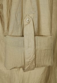Puño de camisa de lino beige con botón, que presenta una textura suave y un diseño plegado, destacado por costuras visibles y detalle del botón.