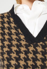 V-kaulusneule, jossa on musta-tan-hakkapeliitto-kuvio, valmistettu pehmeästä neulemateriaalista. Kerroksittain valkoisen napinläpi-paidan päällä.