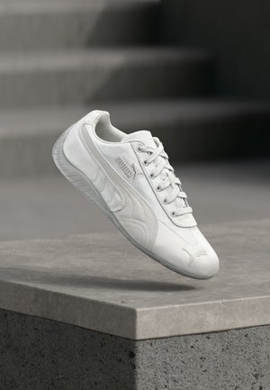 Baskets blanches Puma avec lacets, logo sur le côté et l'avant, flottant au-dessus d'une plateforme en béton grise avec des marches floues en arrière-plan.