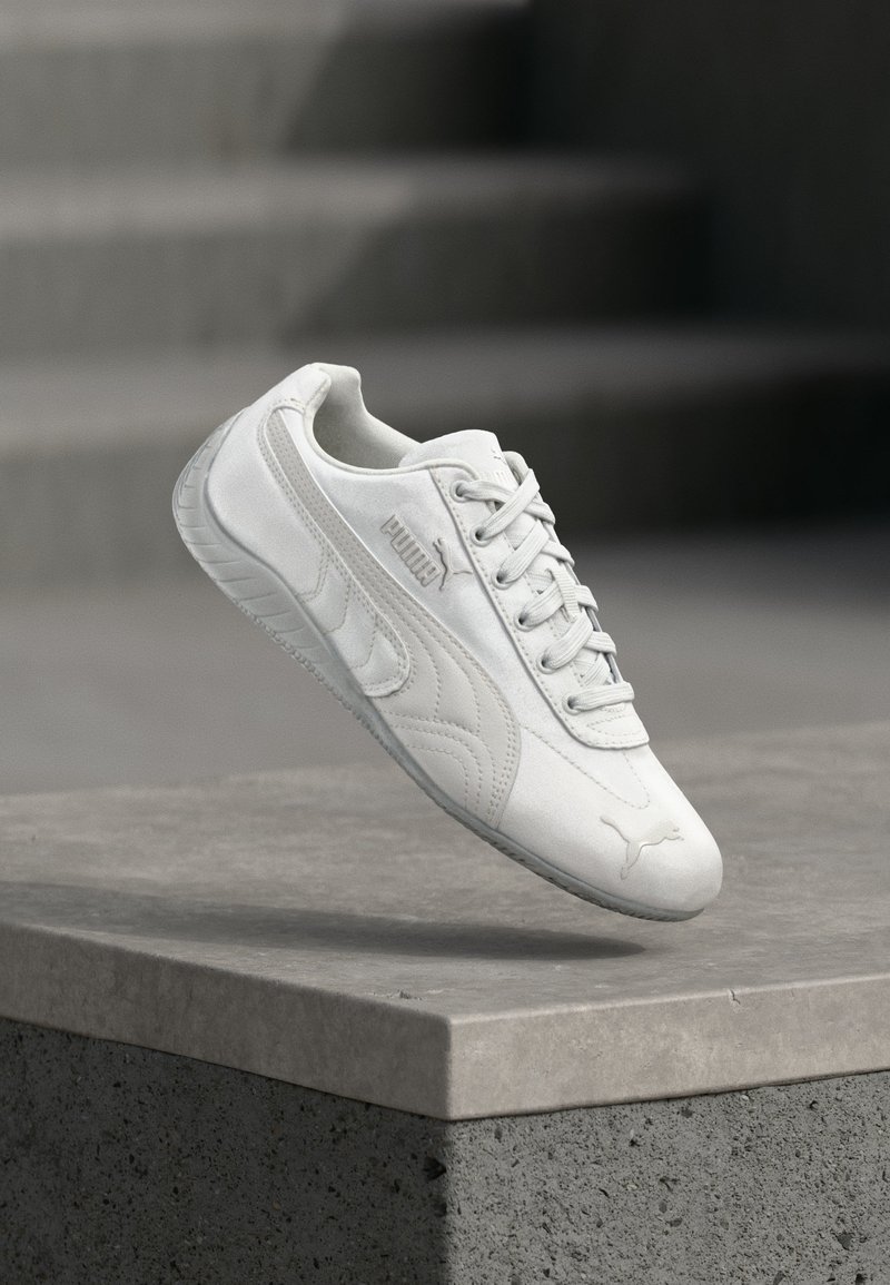 Λευκό αθλητικό παπούτσι Puma με κορδόνια, λογότυπο στο πλάι και στο μπροστινό μέρος, αιωρούμενο πάνω από γκρι τσιμεντένιο βάθρο με θολωμένα σκαλοπάτια στο φόντο.