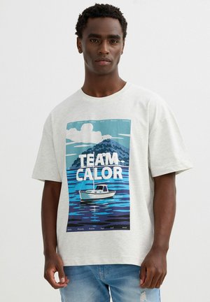 Hombre con camiseta gris claro con gráfico azul de barco y mar y texto "TEAM CALOR", combinado con vaqueros azul claro.