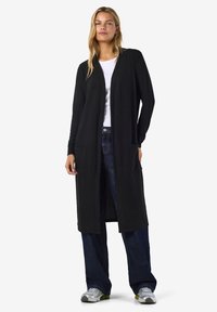 Sort lang cardigan med åpen front, to sidelommer og lange ermer, båret over en hvit skjorte og blå jeans. Glatt stofftekstur.