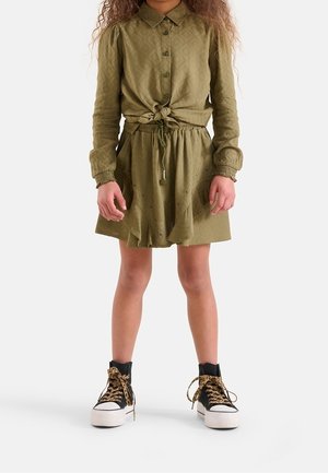 Enfant portant une chemise boutonnée vert olive nouée à la taille, une jupe assortie avec un cordon à pompons, et des baskets montantes noires avec des lacets léopard.