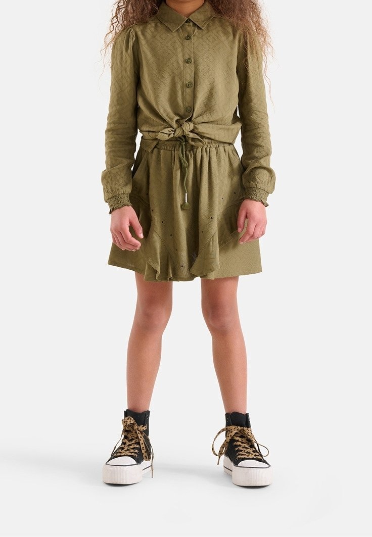 Enfant portant une chemise boutonnée vert olive nouée à la taille, une jupe assortie avec un cordon à pompons, et des baskets montantes noires avec des lacets léopard.