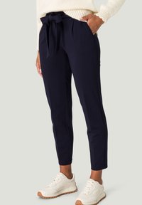 Femme portant un pantalon tapered bleu marine avec une ceinture nouée à la taille, associé à des baskets blanches chunky et un pull en tricot crème.